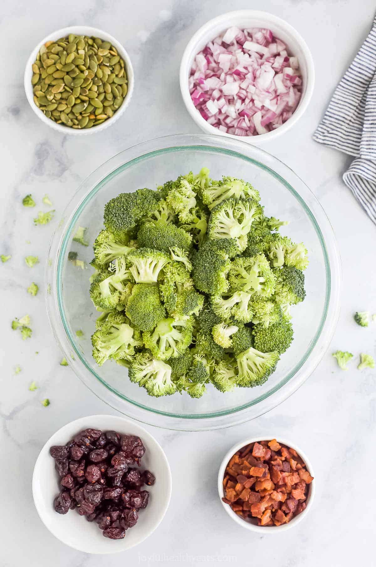 A bowl of broccoli florets beside a bowl of diced onions along with the remaining salad ingredients on a marble surface