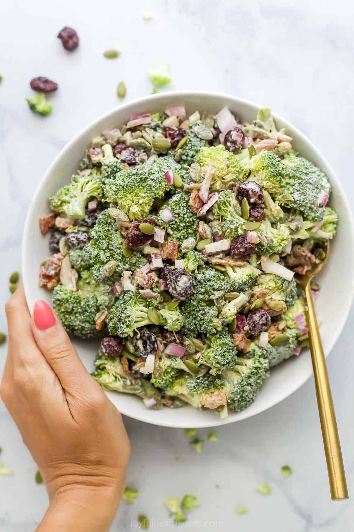 A hand holding a big bowl of broccoli salad with a gold-colored spoon inside
