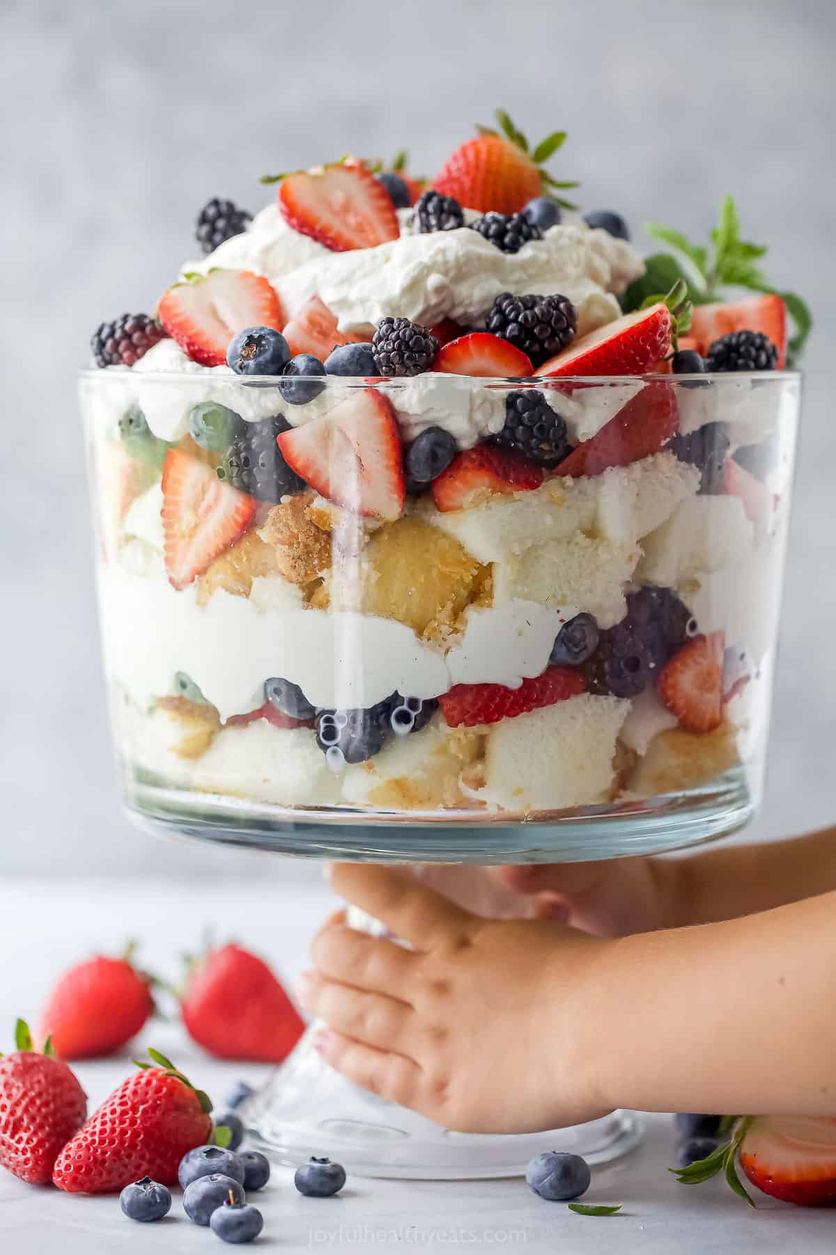 A strawberry shortcake trifle in a large glass trifle dish with a pair of hands holding the base