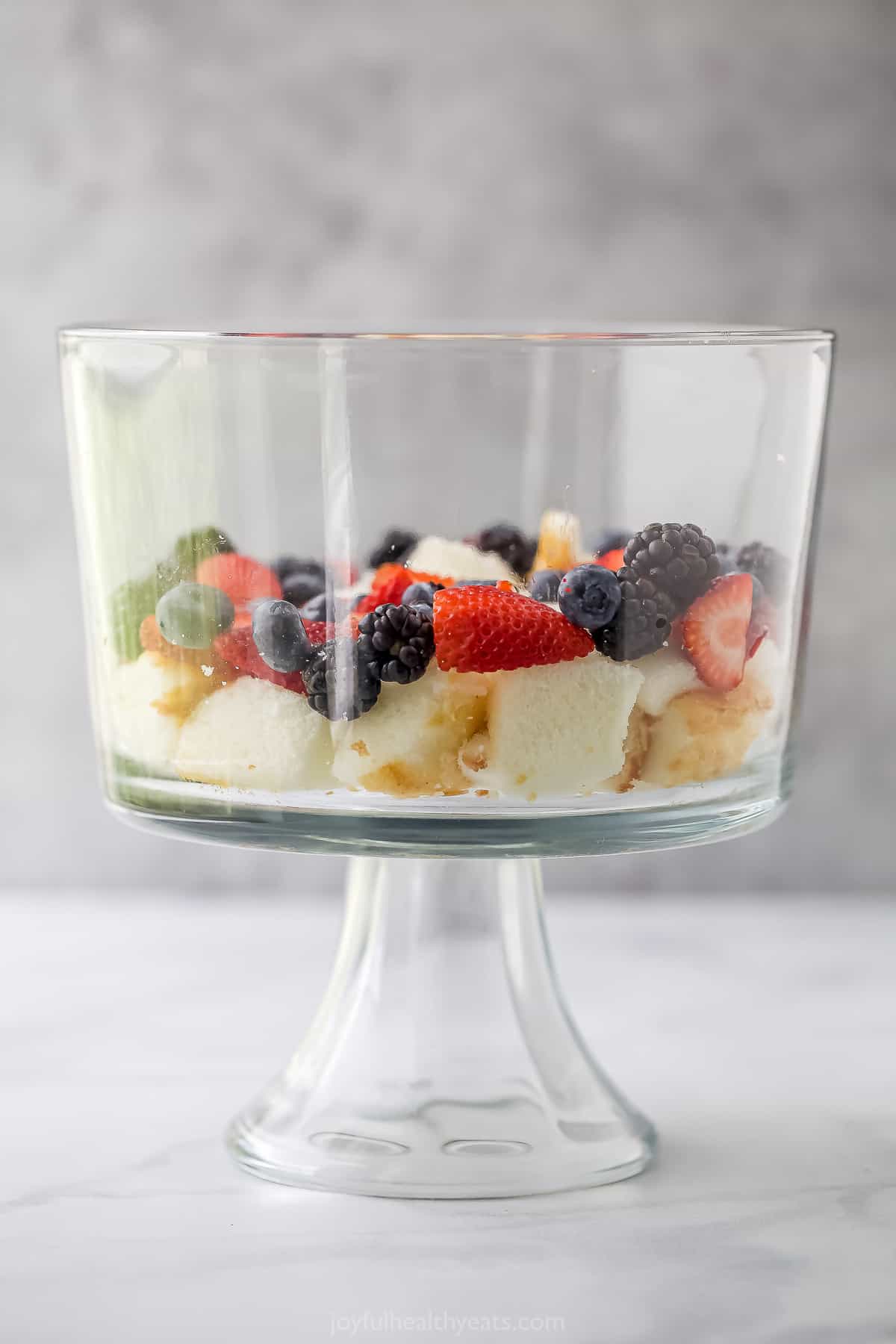Layers of cubed angel food cake, whipped cream and fresh berries in a large glass dish with a glass base