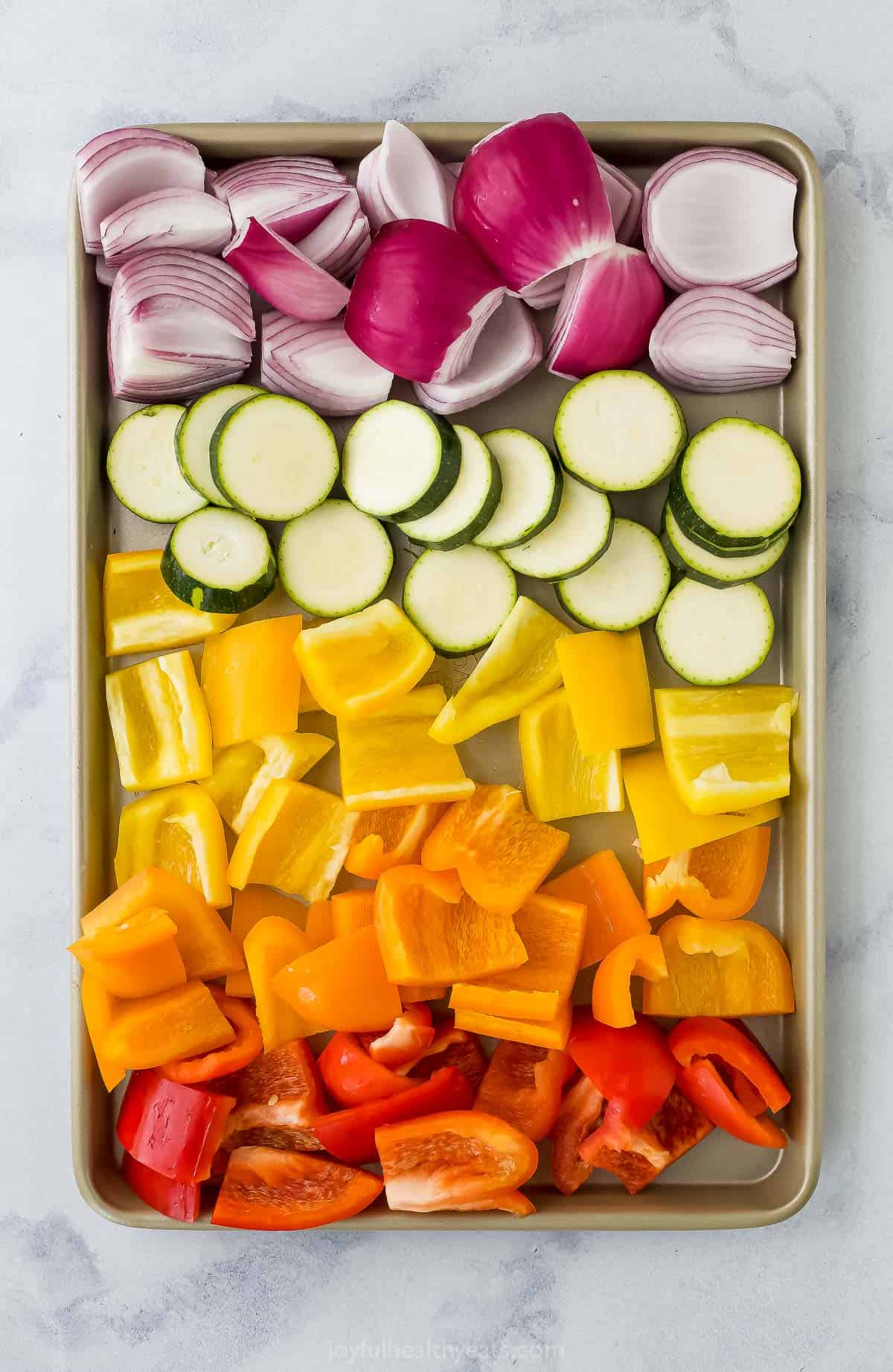 Chopped peppers on a baking sheet with quartered red onions and zucchini coins