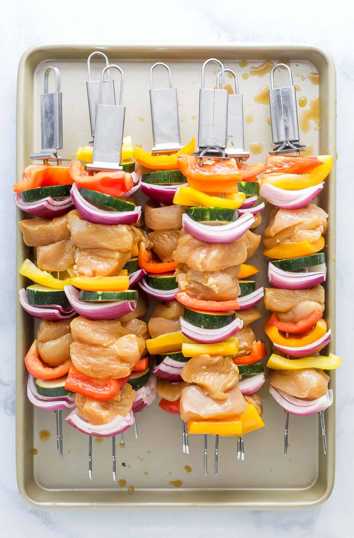 Raw chicken skewers piled onto a baking sheet with a raised rim