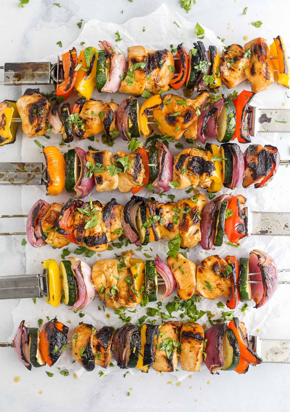 Six marinated chicken kabobs on a marble countertop with a sheet of parchment paper underneath them