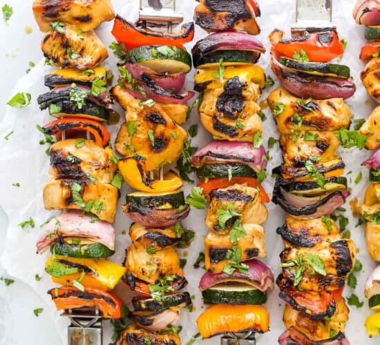 Four grilled chicken kabobs lined up on a countertop with a fifth one partially in frame