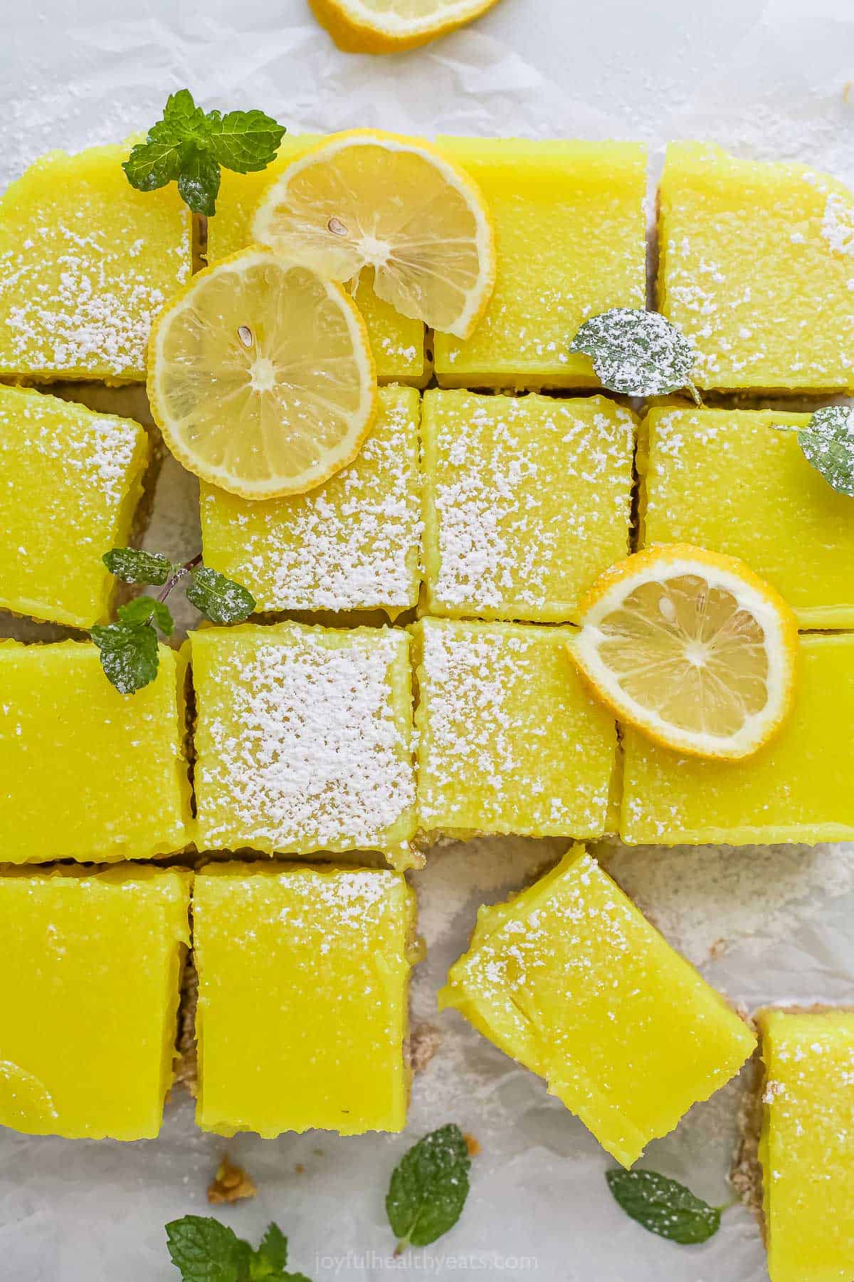 Sixteen vegan lemon bars on a sheet of parchment paper with a little powdered sugar sprinkled over them