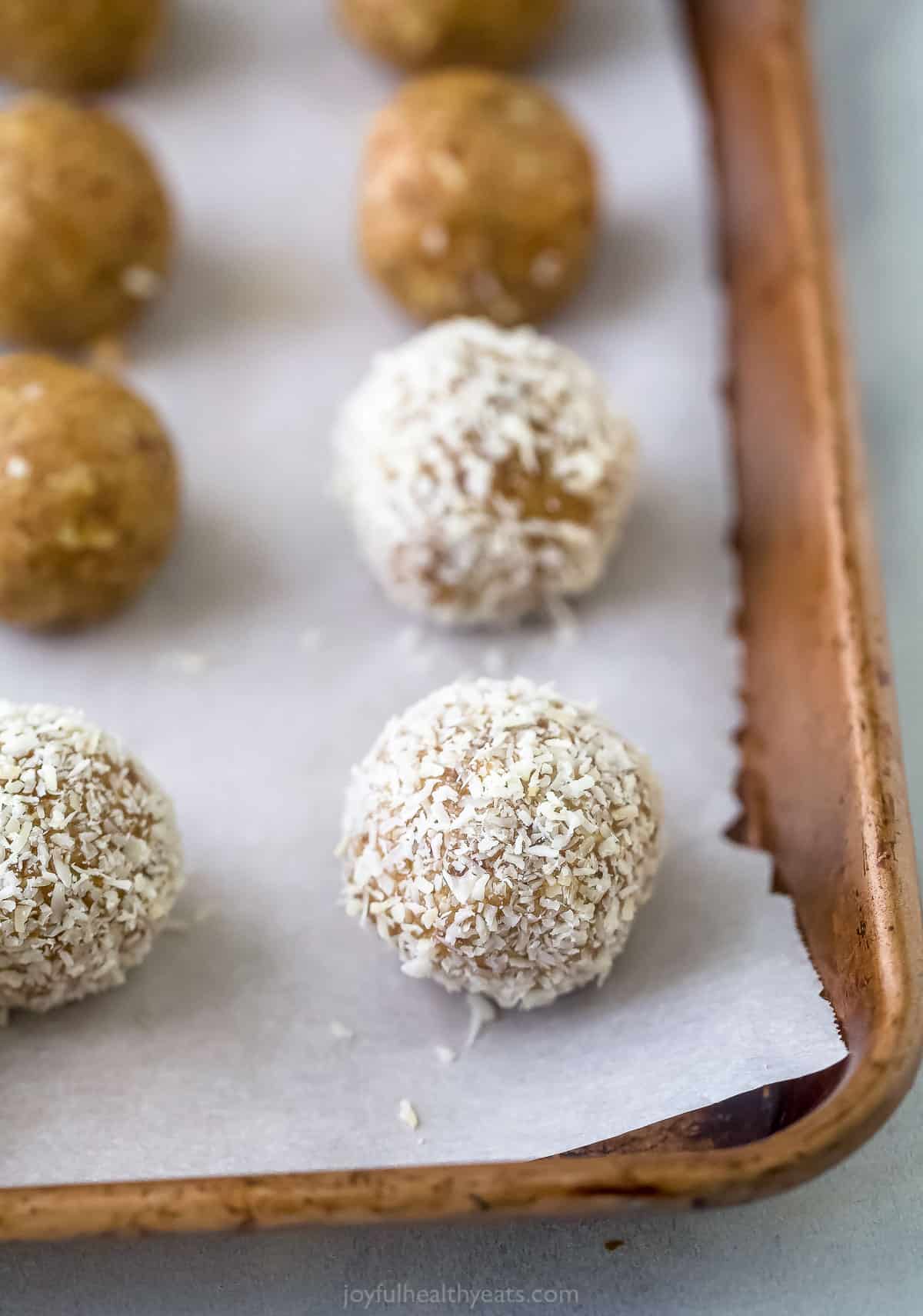 Energy balls on a parchment-lined baking sheet with some of them yet to be coated in shredded coconut