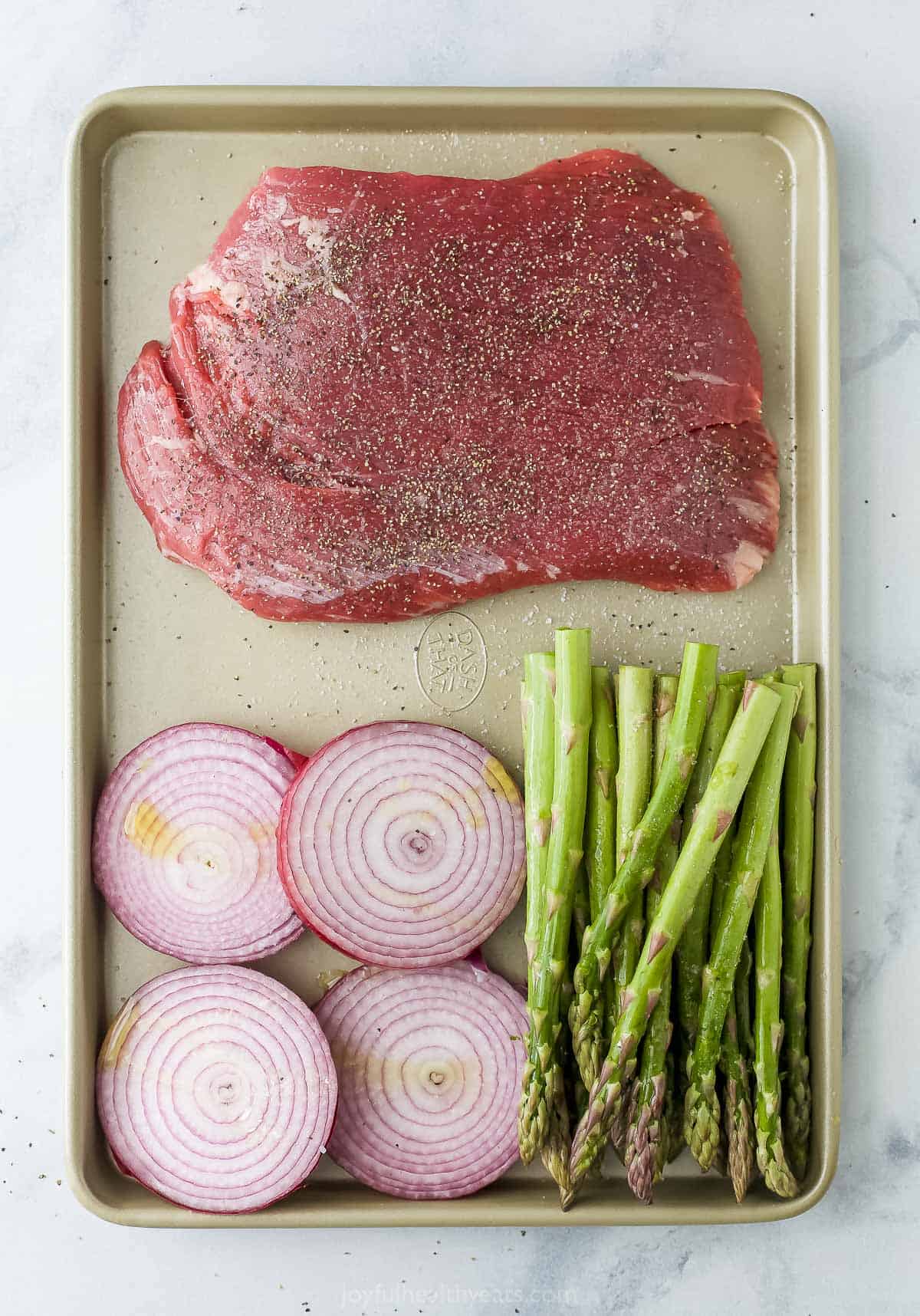 Raw beef on a cookie sheet with sliced red onions and fresh asparagus spears