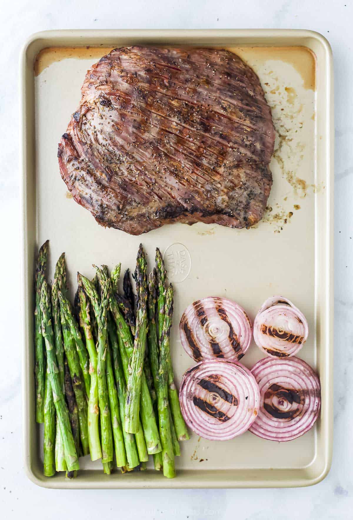 Grilled beef on a baking sheet beside grilled asparagus and grilled red onion