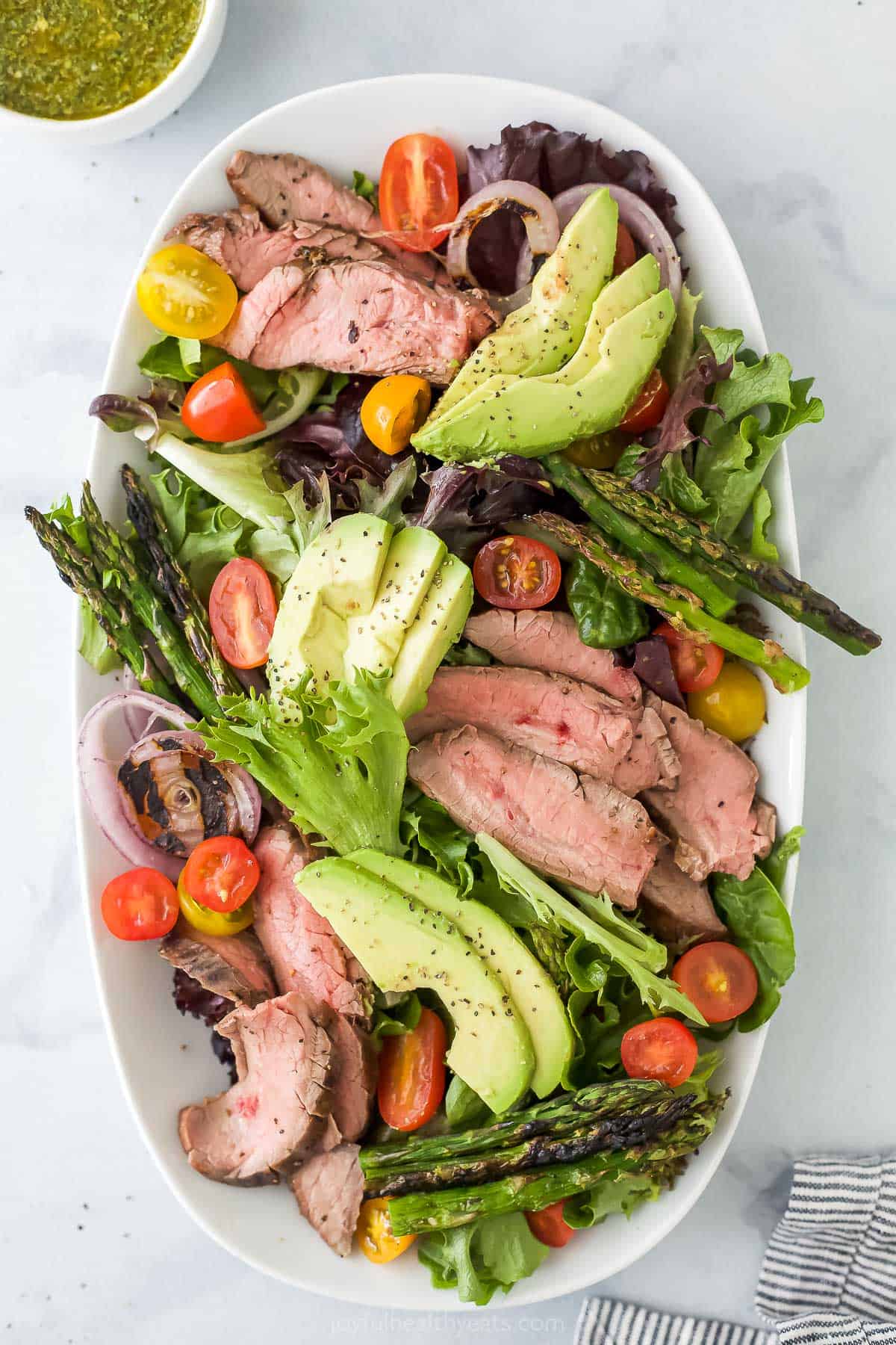 A platter full of steak salad with a dish of chimichurri and a kitchen towel beside it