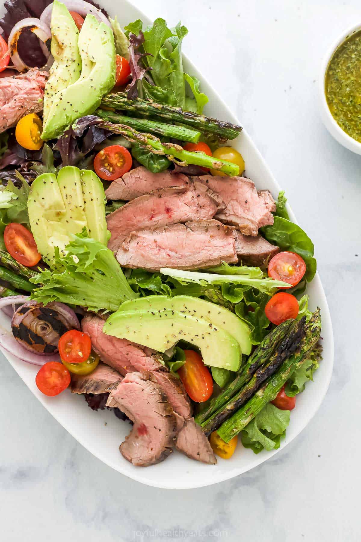 A grilled flank steak salad on a serving platter beside a dish of chimichurri