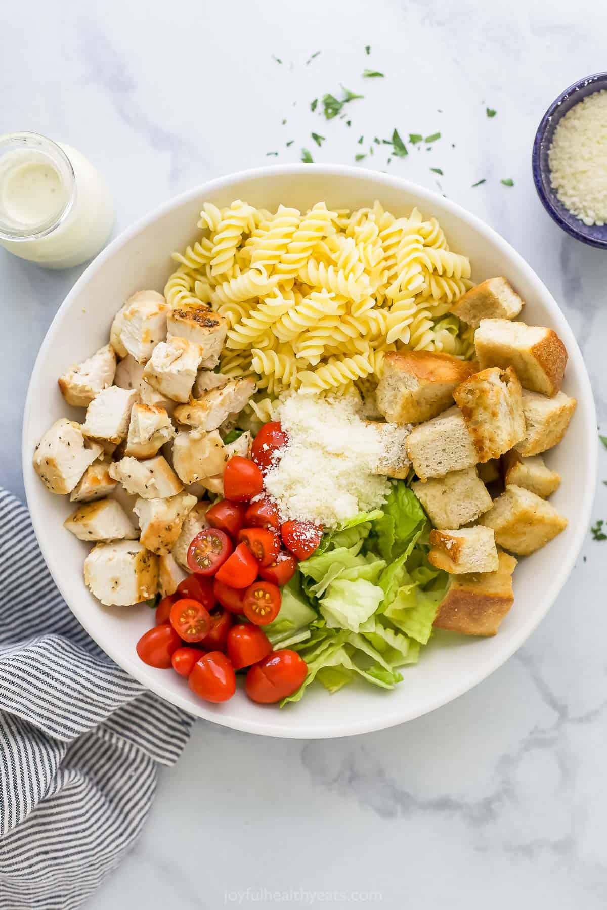 The garlic herb croutons and all of the salad ingredients inside of a large mixing bowl beside a striped dishtowel