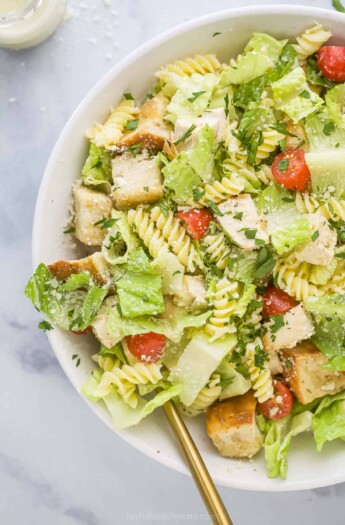 A chicken caesar pasta salad in a big white bowl with a bottle of homemade caesar dressing beside it