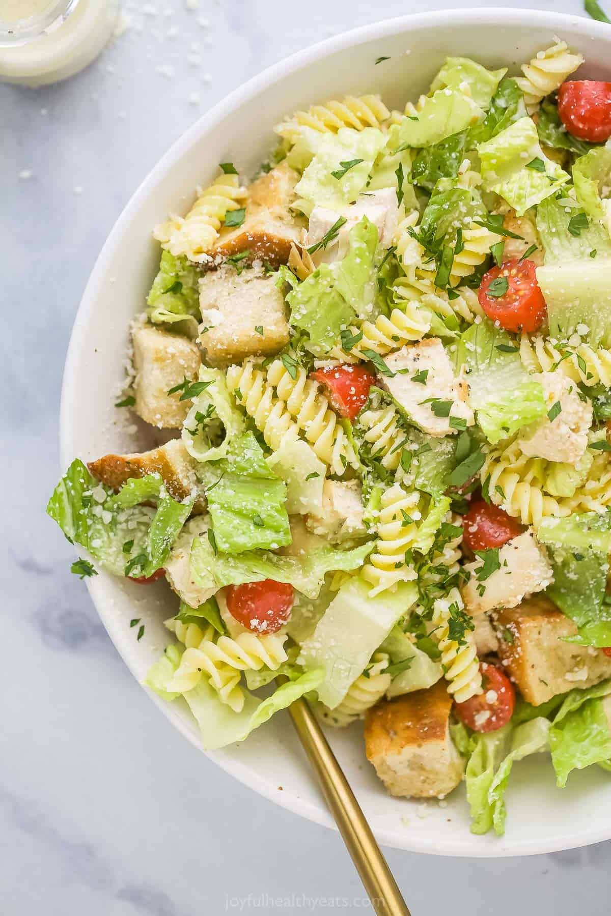 A chicken caesar pasta salad in a big white bowl with a bottle of homemade caesar dressing beside it