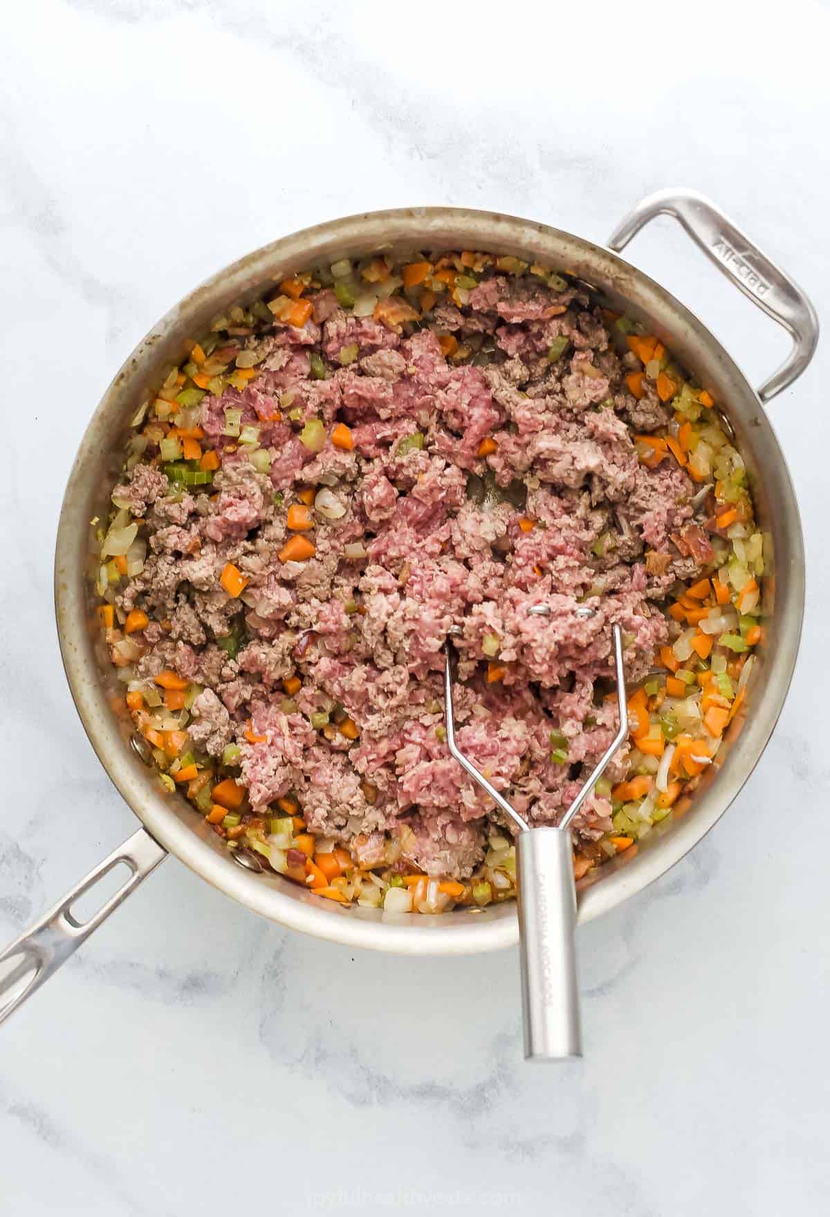 Saute pan with ground beef being mashed up and mixed with cooked vegetables