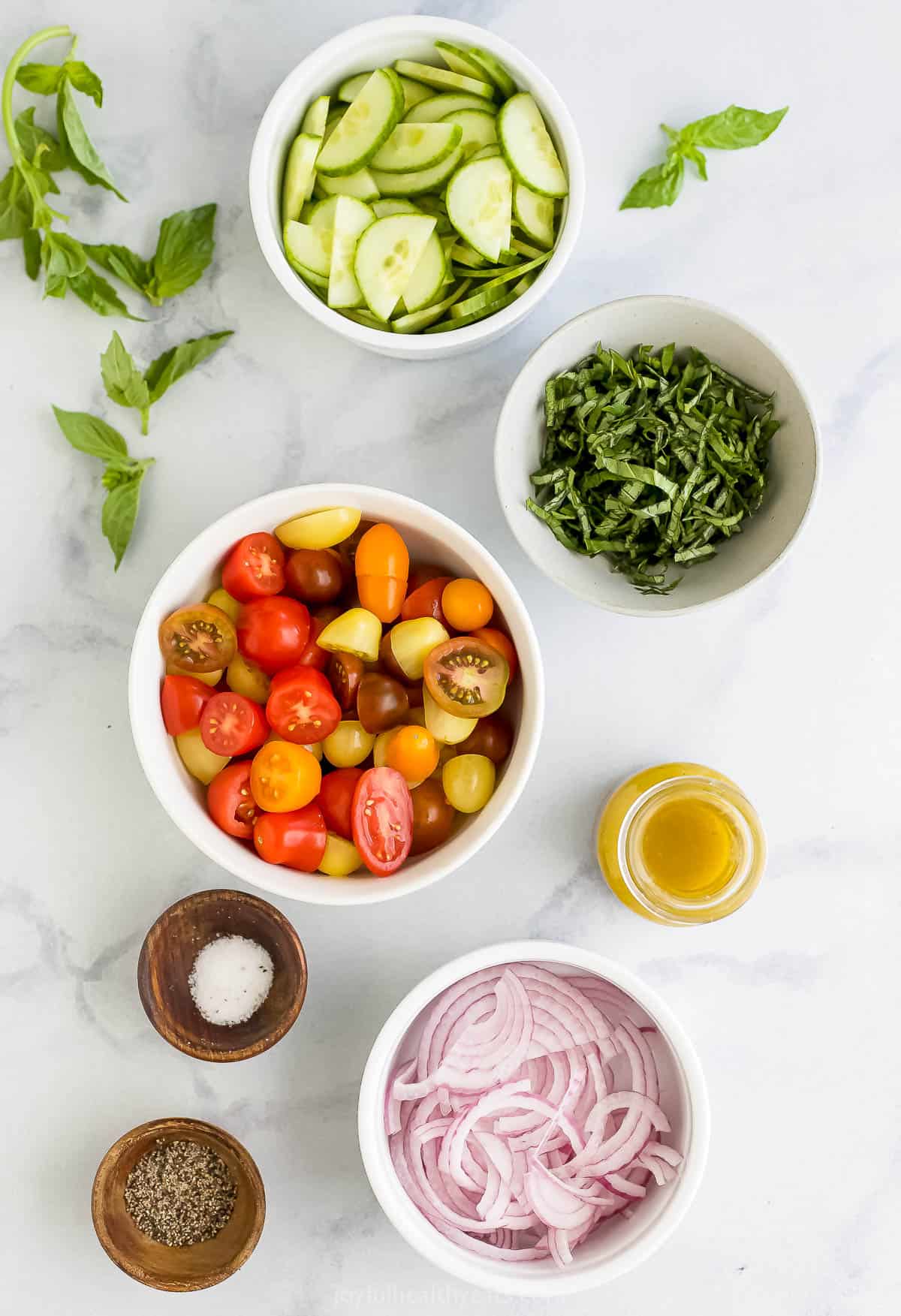 A bowl of sliced cucumbers, a bowl of halved cherry tomatoes and the rest of the panzanella ingredients arranged on a kitchen counter