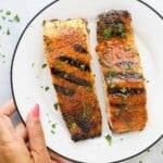 Two grilled salmon fillets on a plate with a hand grabbing it