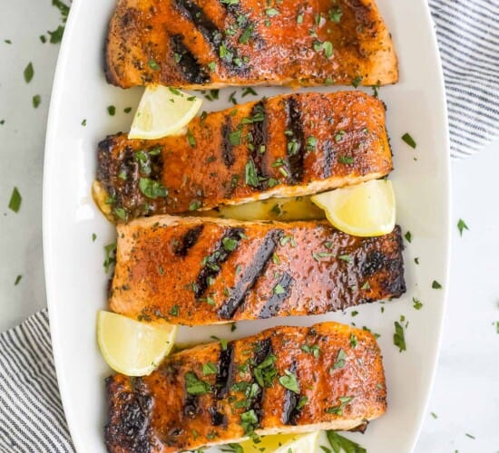A white platter holding four blackened salmon fillets and five fresh lemon wedges