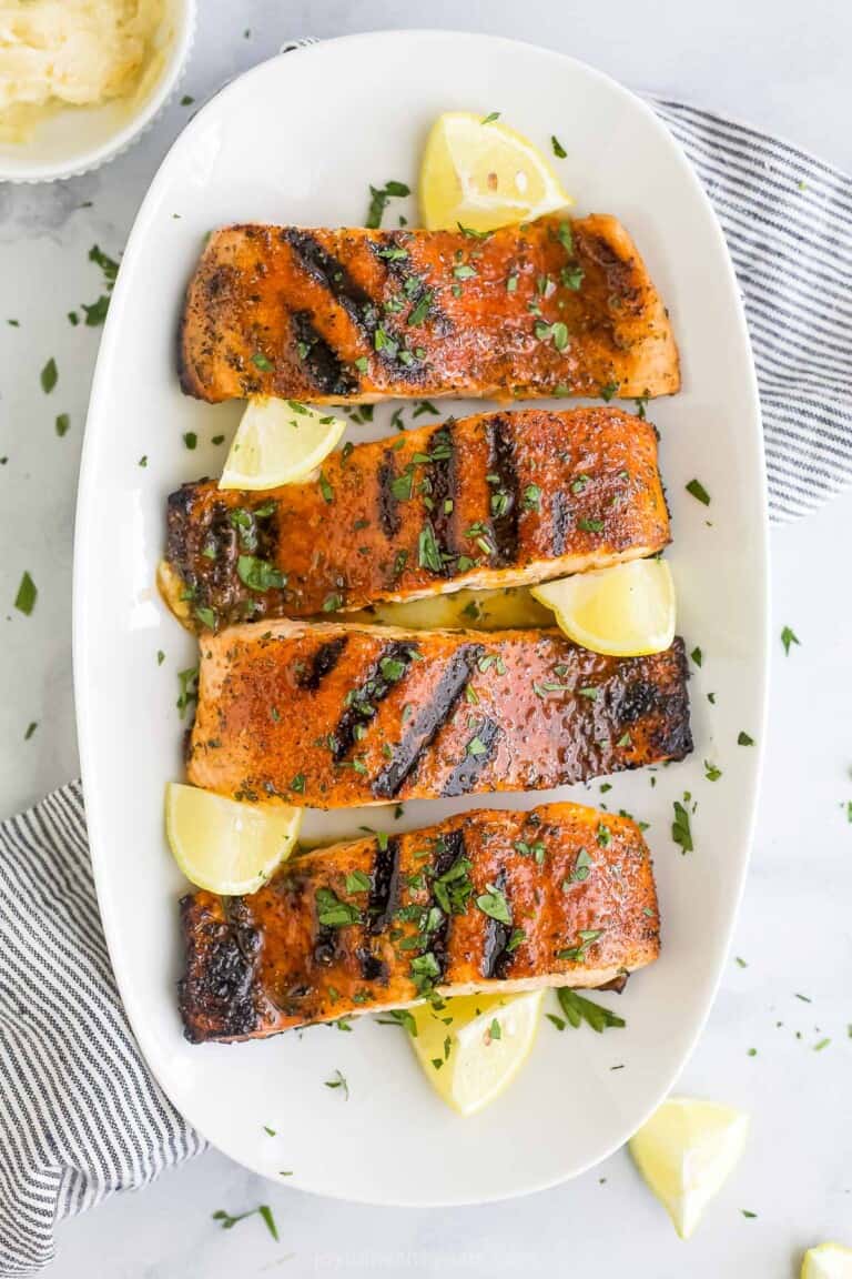 A white platter holding four blackened salmon fillets and five fresh lemon wedges