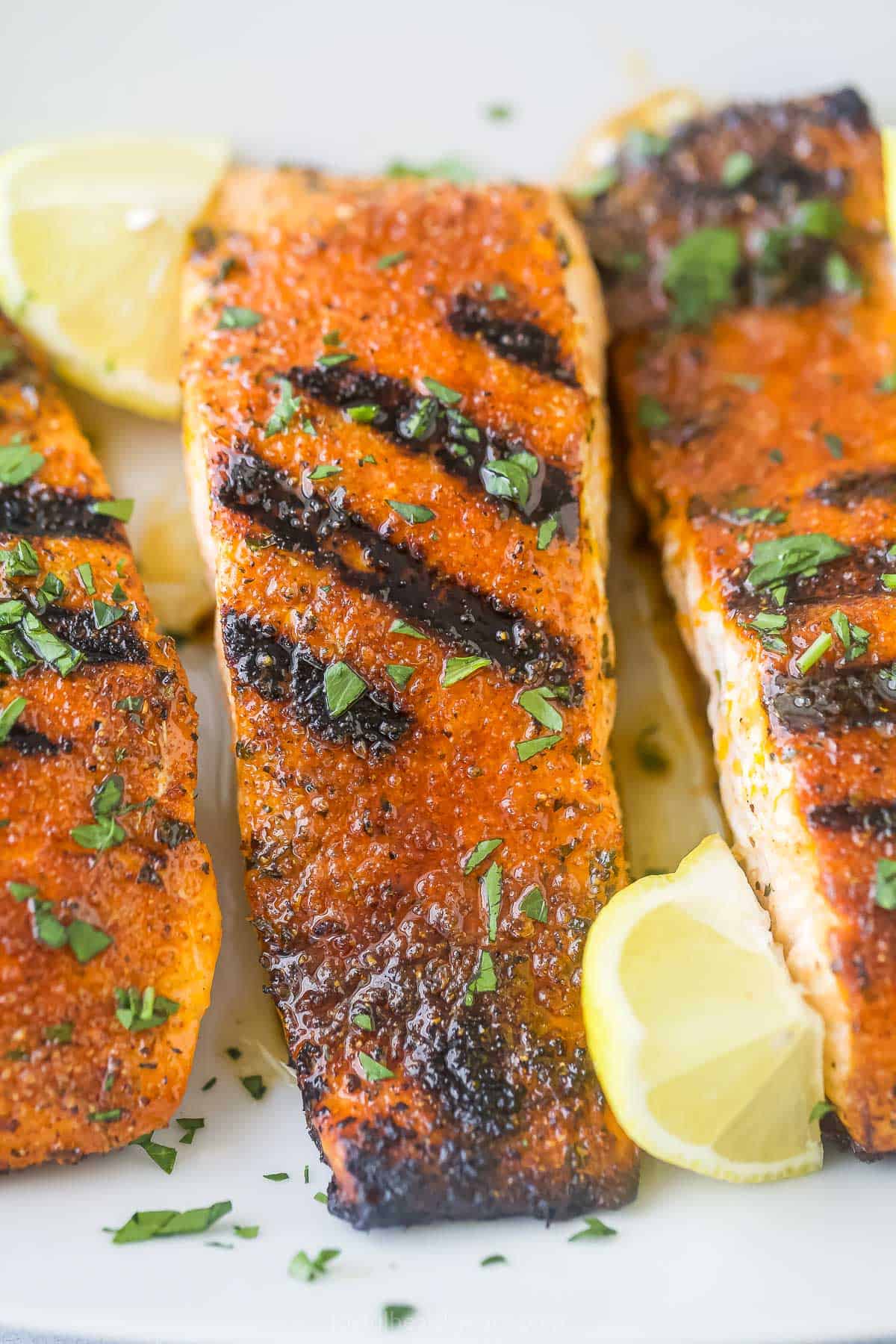 A close-up shot of three blackened salmon fillets on a plate with fresh lemon wedges.