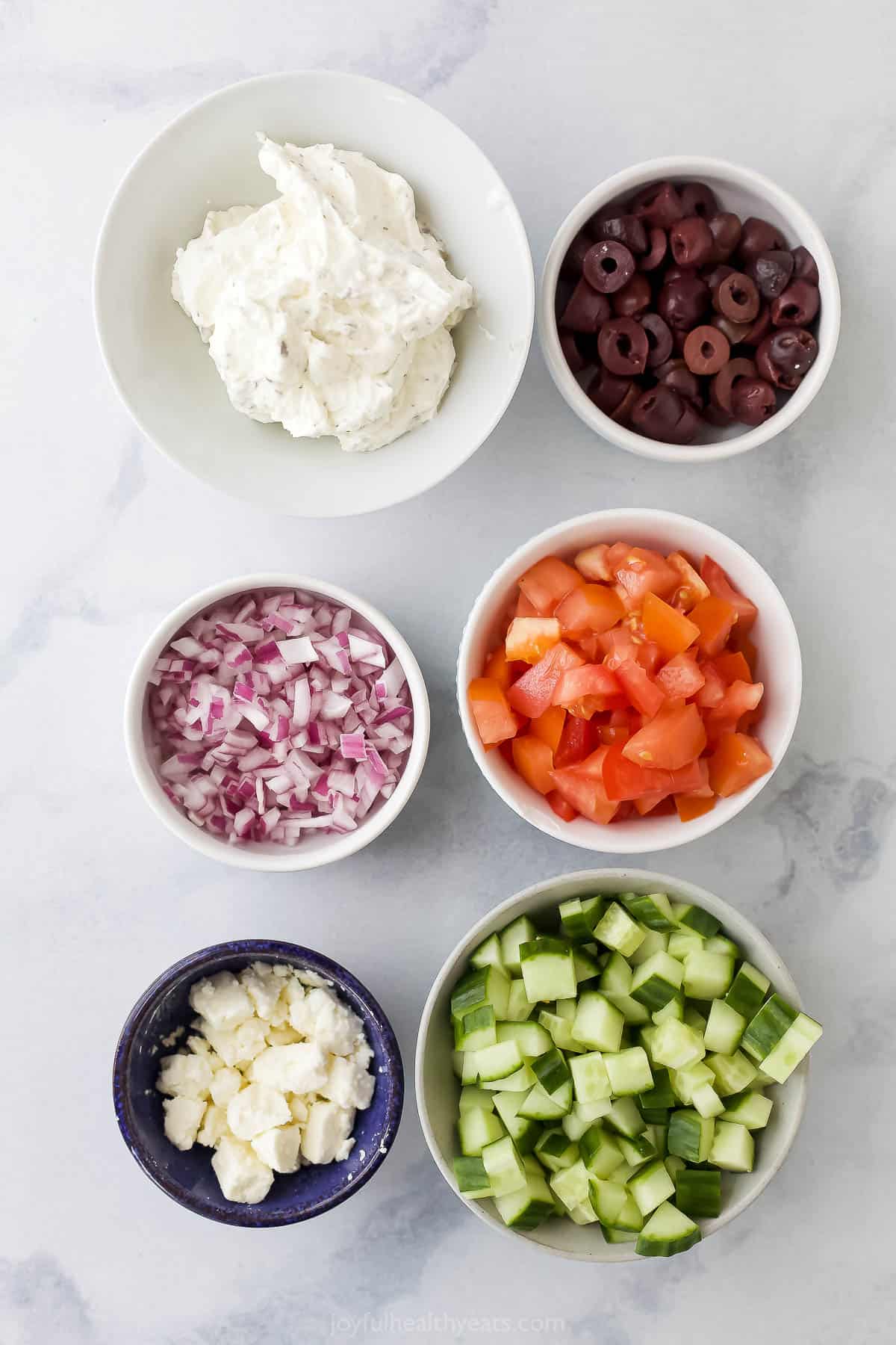 Halved kalamata olives, diced red onion, crumbled feta and the rest of the dip ingredients in separate bowls on a marble surface