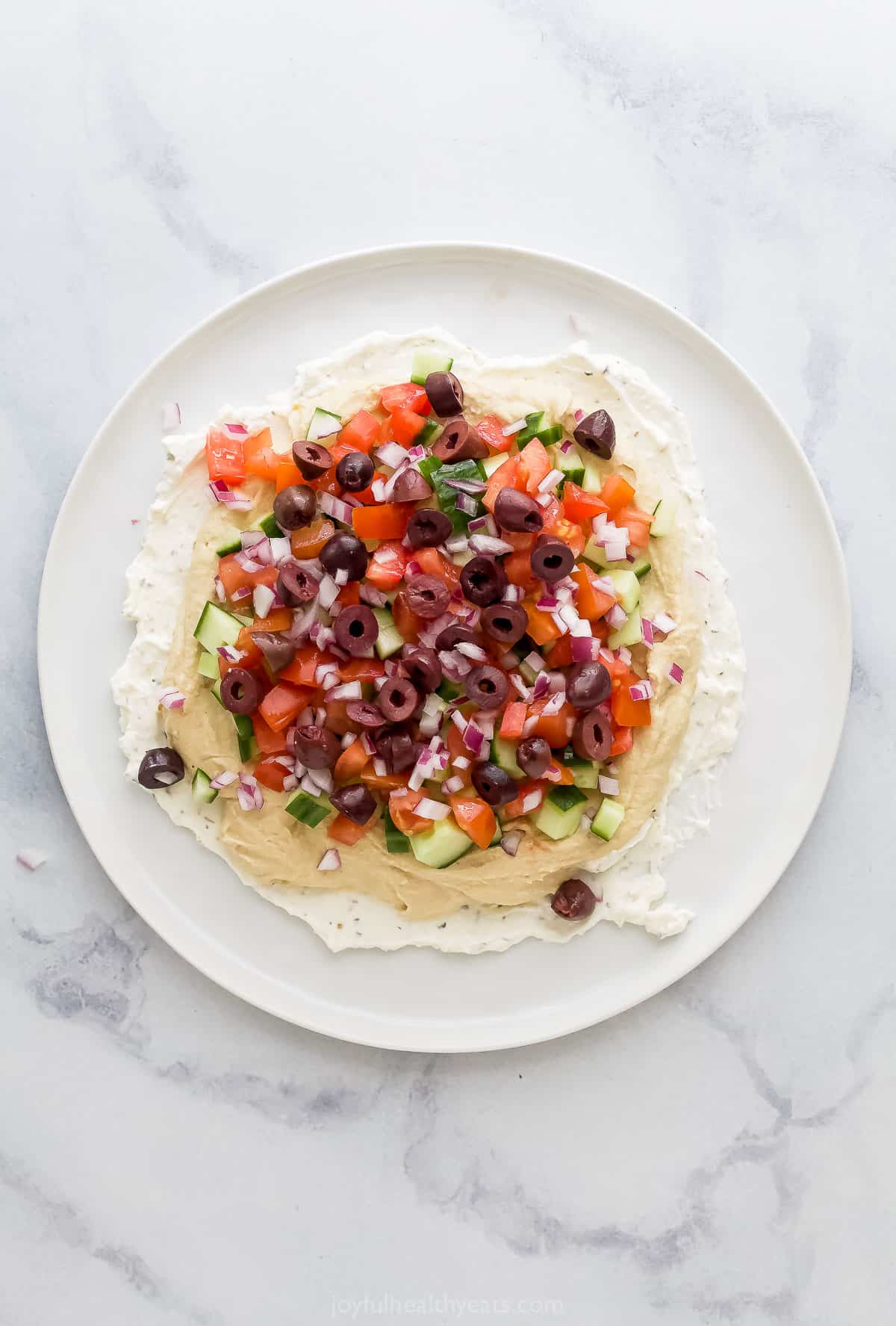 Halved olives, diced tomatoes, diced red onion and diced cucumber layered over hummus and whipped feta on a plate