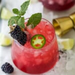 A spicy blackberry margarita in a short cocktail glass with fresh fruit surrounding it