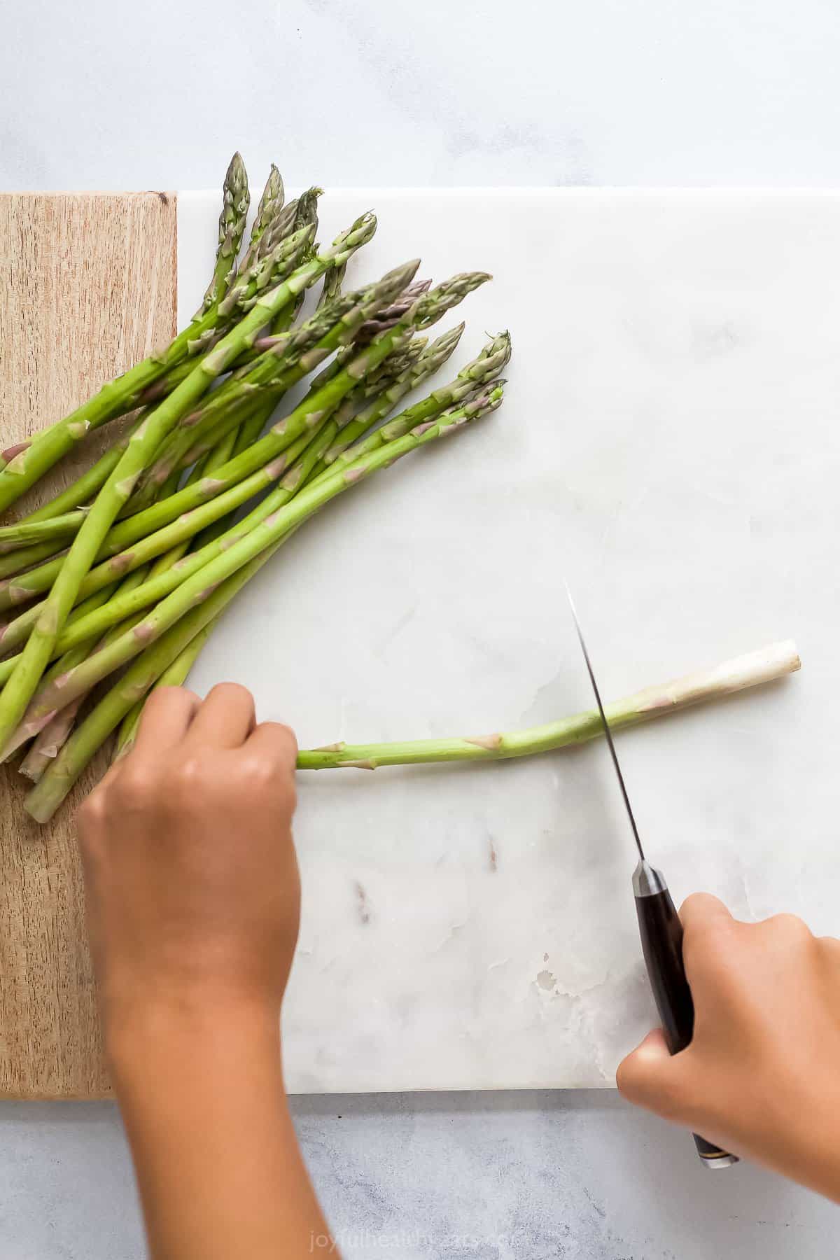 A sharp knife being used to cut the woody stem off a stalk of asparagus over a cutting board