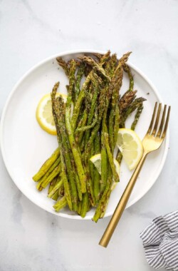 A plate full of roasted asparagus with three lemon slices and a fork beside the veggies