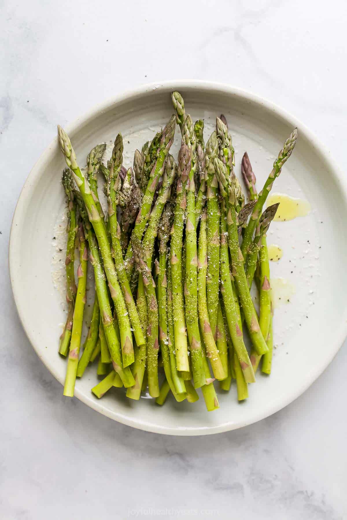 Raw asparagus stalks piled onto a plate with oil, garlic powder, salt and pepper sprinkled over them