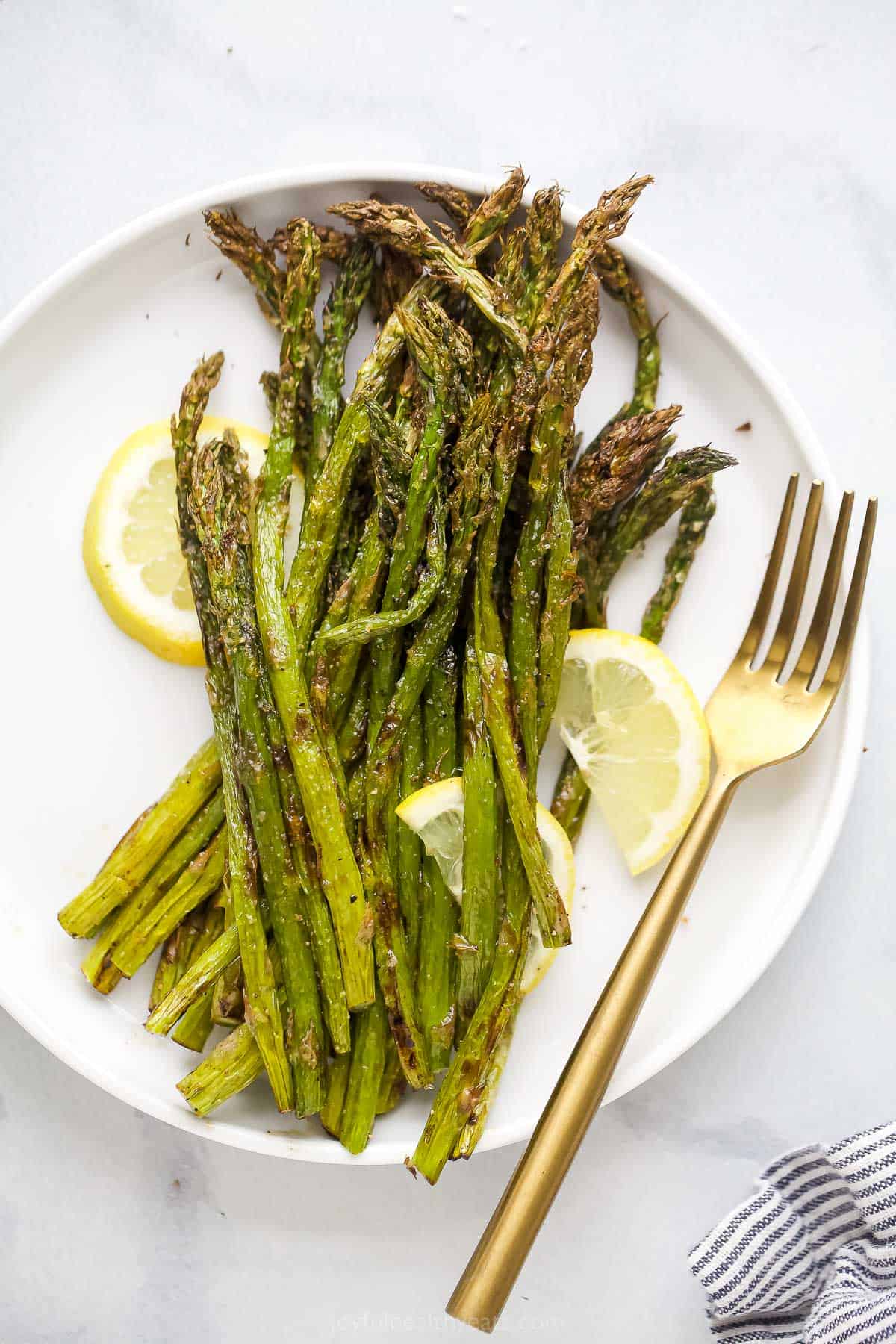 Air-fried asparagus on a white plate with fresh lemon slices and a golden fork