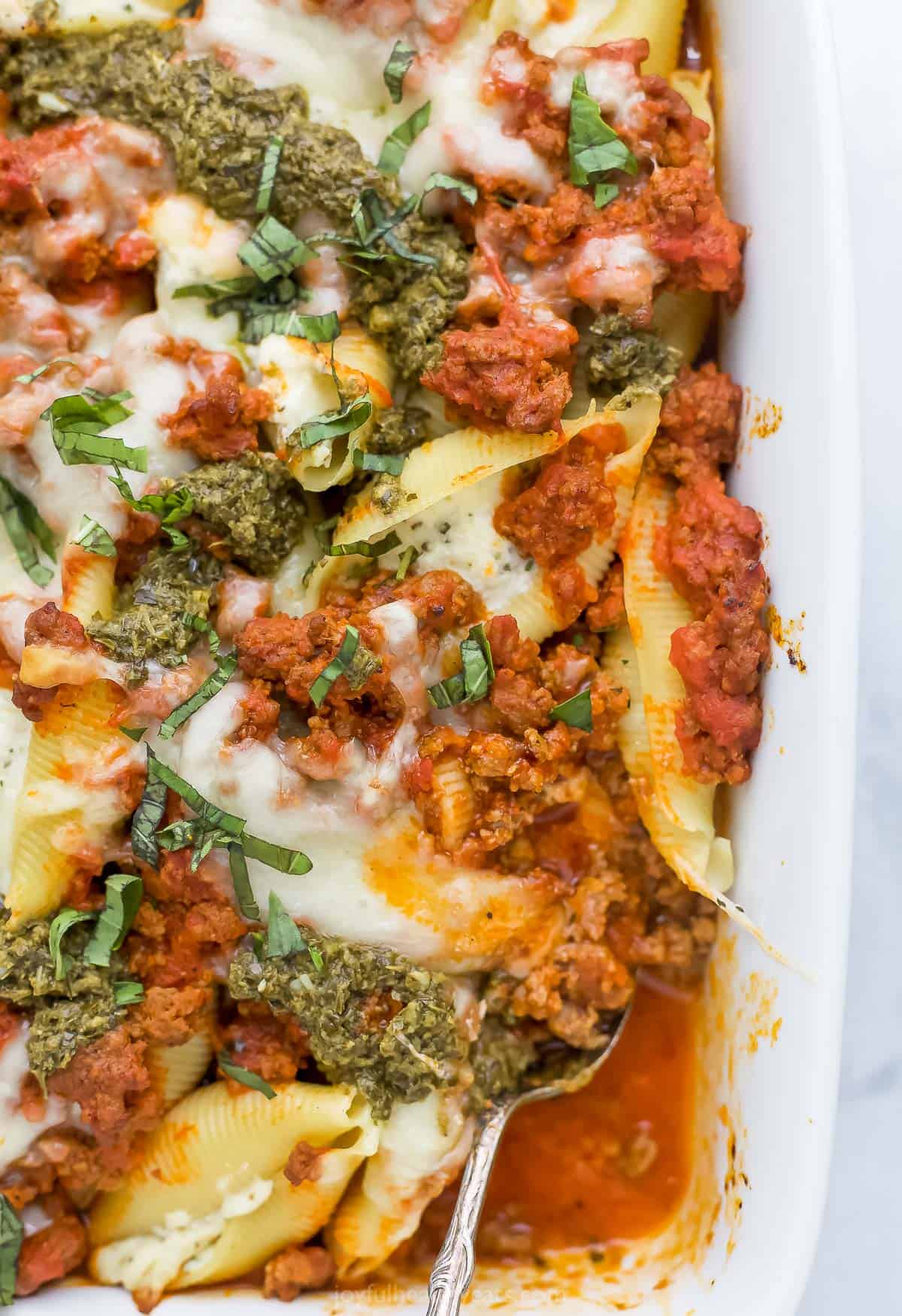 A white casserole dish containing stuffed shells with meat sauce and a garnish of pesto and fresh basil