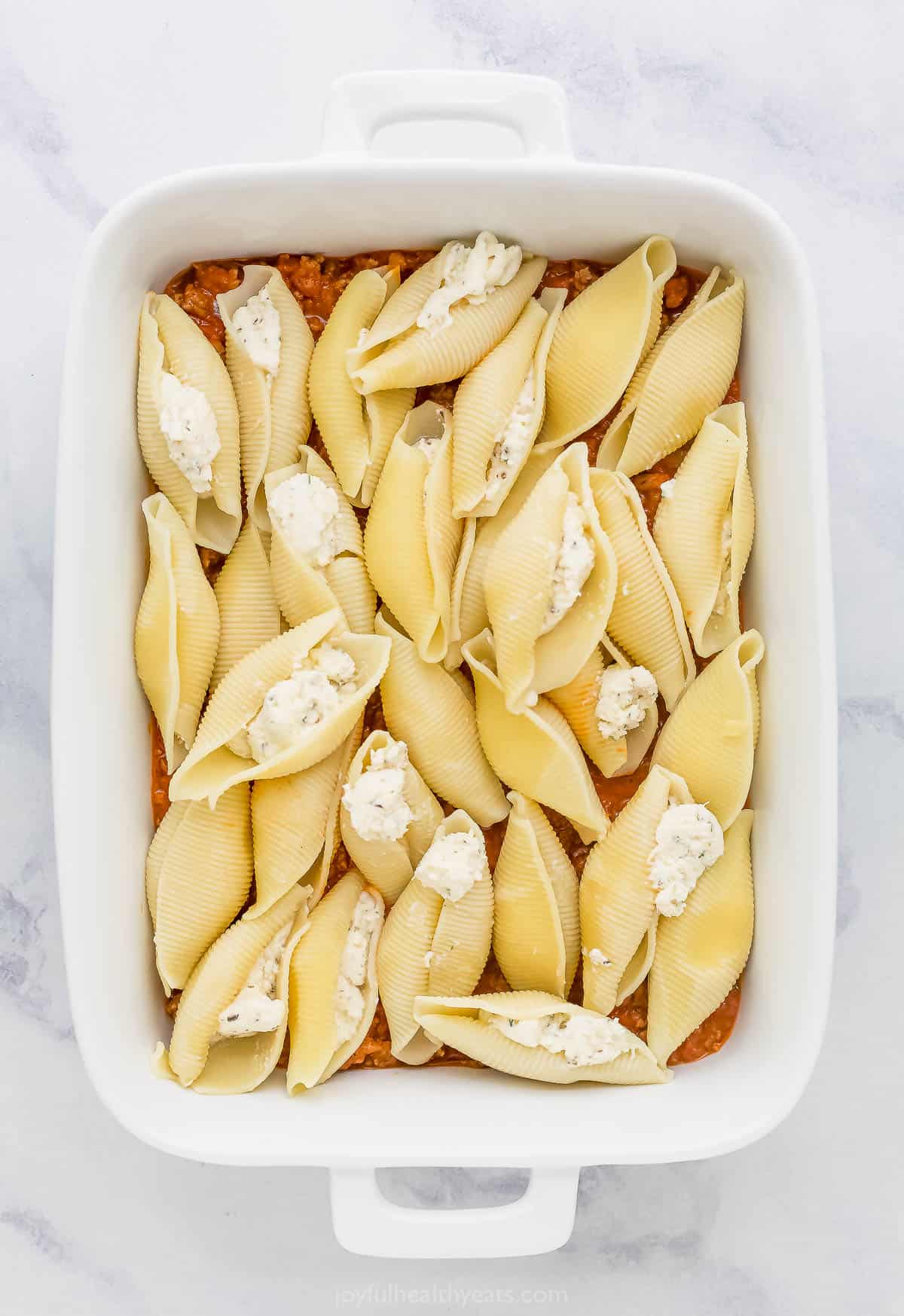 Meat sauce and stuffed shells layered into a baking dish on a marble countertop