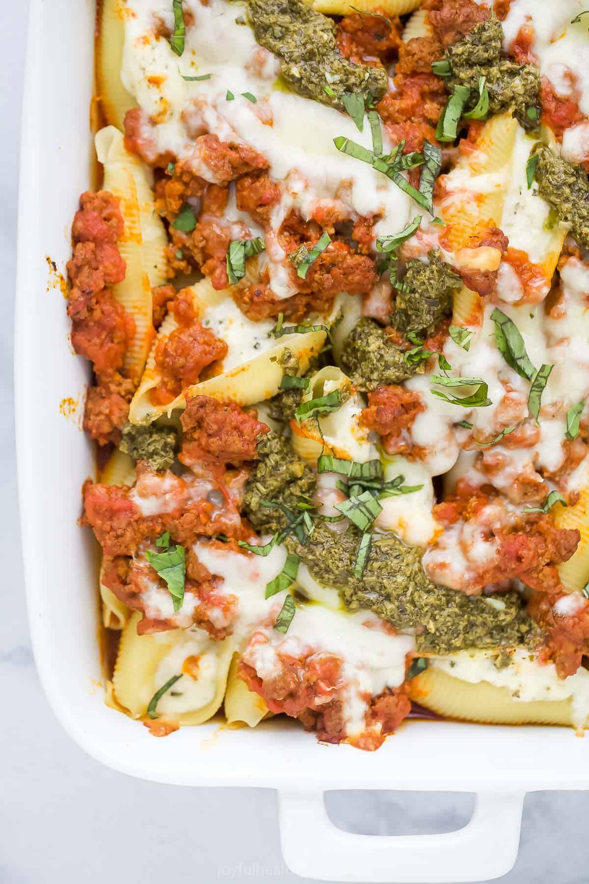 A white baking dish containing cheese stuffed shells with meat sauce and a drizzle of pesto