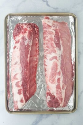 two racks of uncooked ribs on a foil lined sheet tray