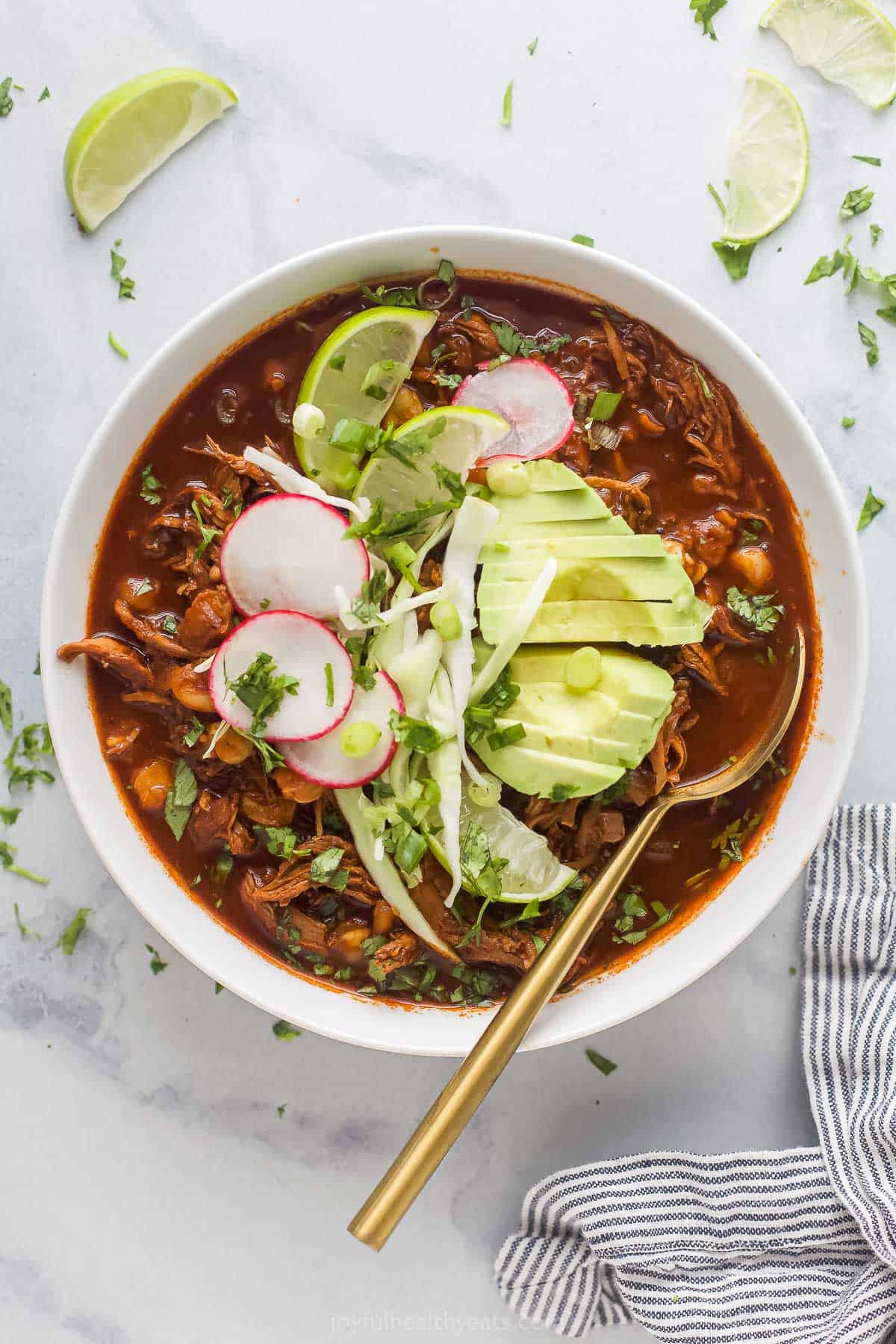 a bowl of red sauce based chicken stew garnished with radishes, lime wedges, cilantro, and avocado slices
