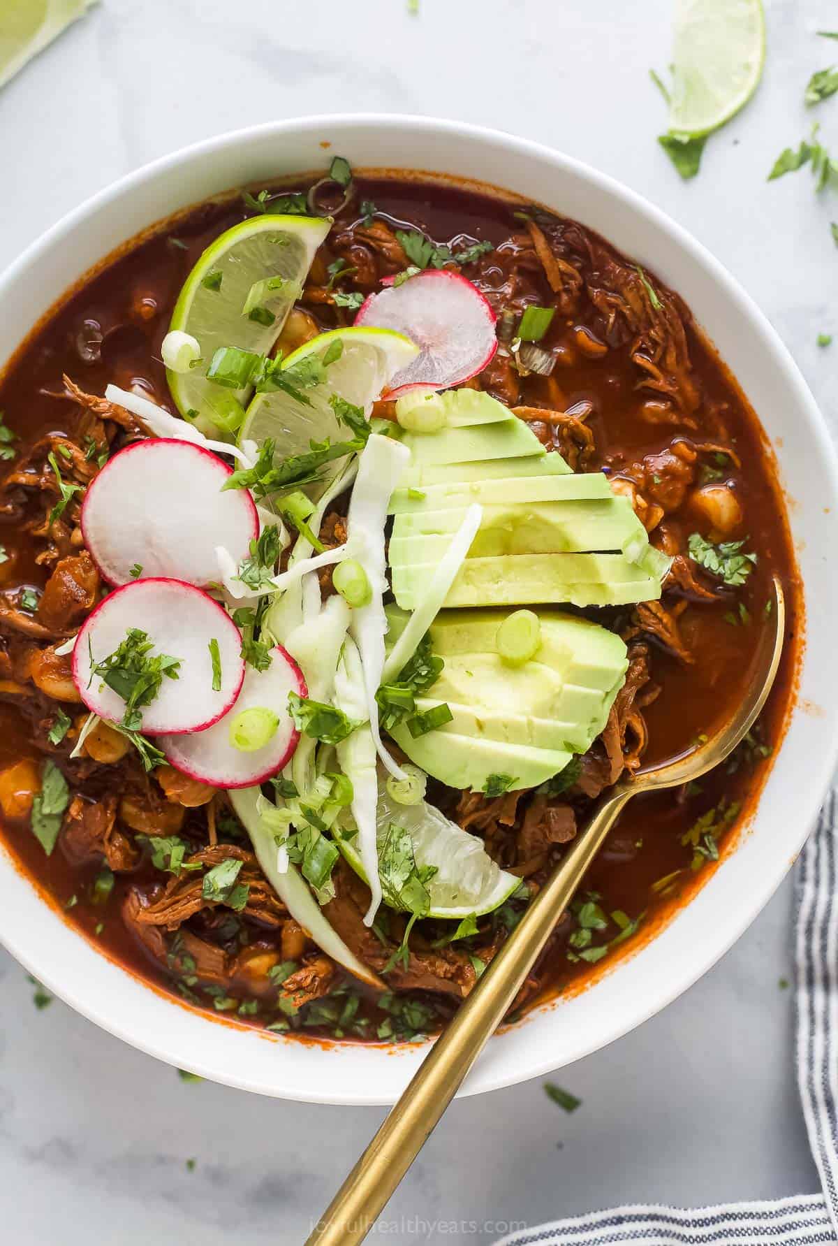 a bowl of red sauce based chicken stew garnished with radishes, lime wedges, cilantro, and avocado slices