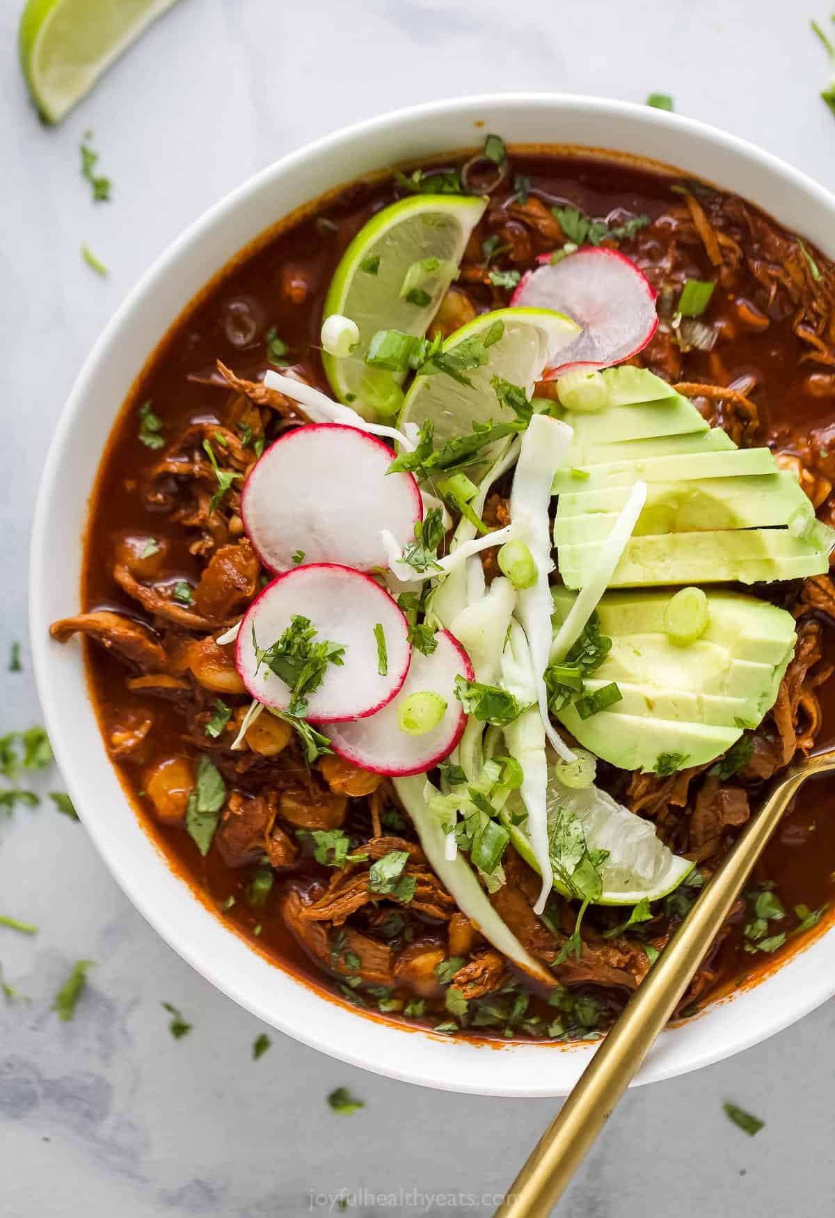 a bowl of red sauce based chicken stew garnished with radishes, lime wedges, cilantro, and avocado slices