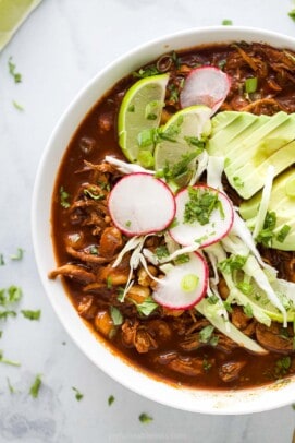 a bowl of red sauce based chicken stew garnished with radishes, lime wedges, cilantro, and avocado slices