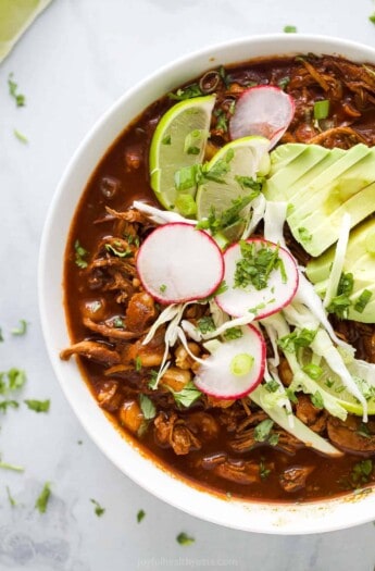 a bowl of red sauce based chicken stew garnished with radishes, lime wedges, cilantro, and avocado slices