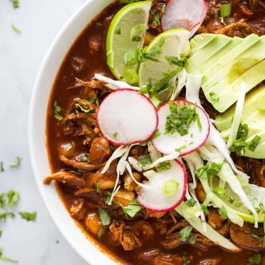 a bowl of red sauce based chicken stew garnished with radishes, lime wedges, cilantro, and avocado slices