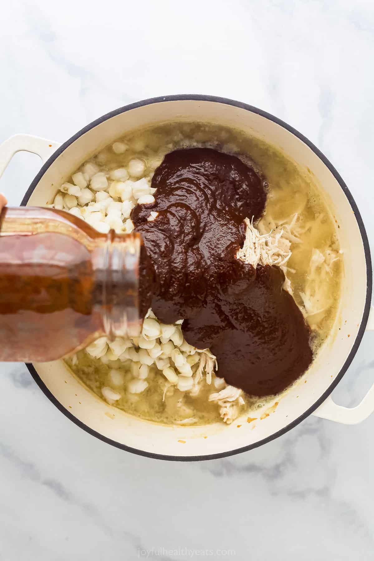 pouring red enchilada sauce into a pot with chicken, broth, and hominy