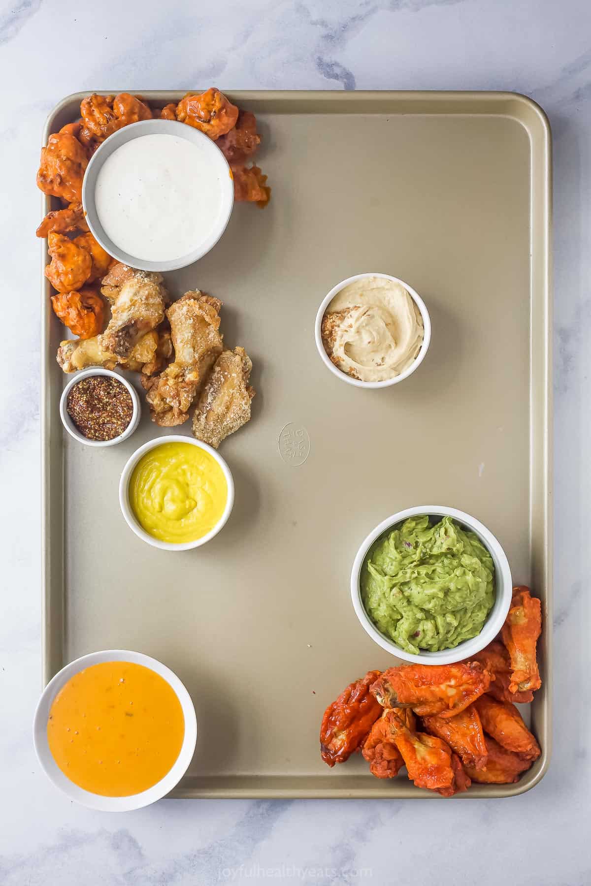 building a snack tray with a sheet tray filled with small bowls with various dips and a portion of the tray being covered with chicken wings