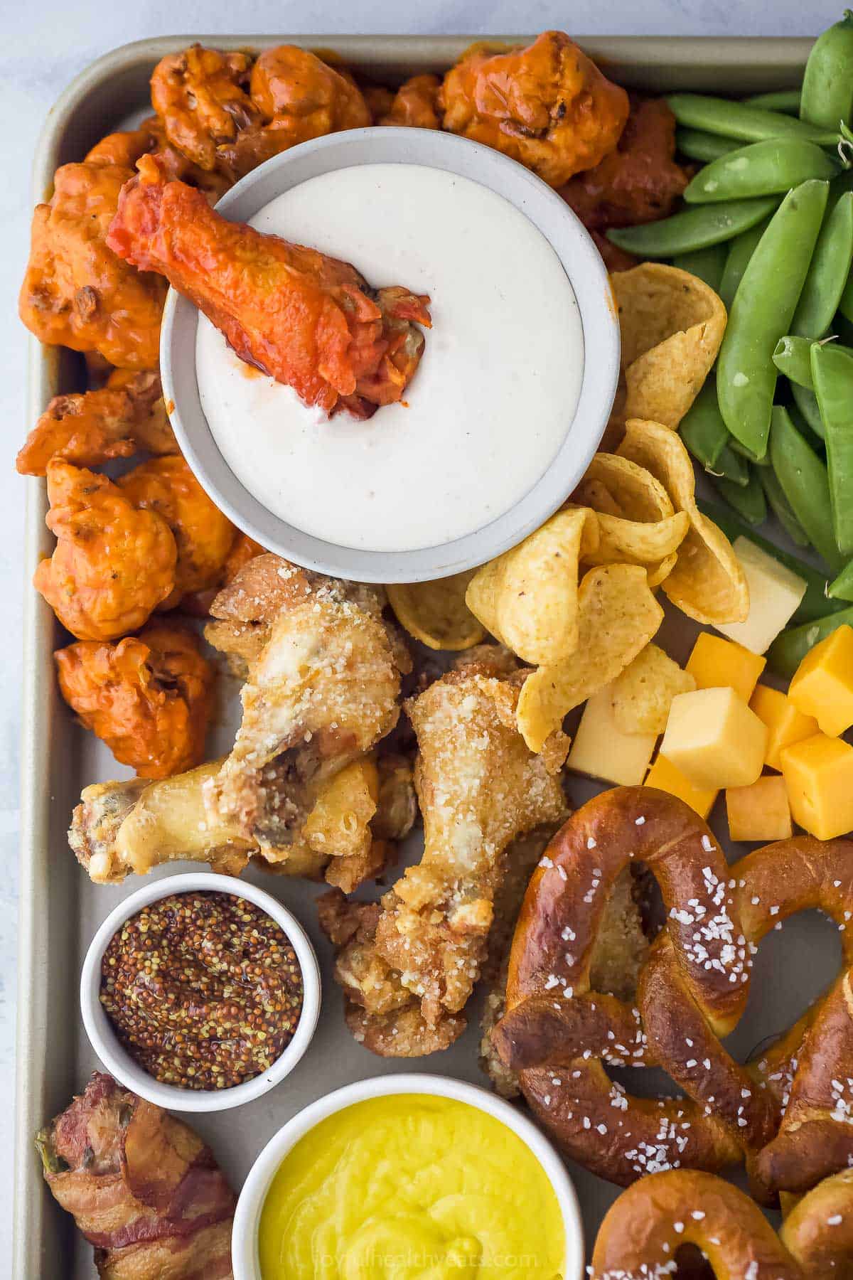 an assortment of snack foods like chicken wings, pretzels, cheese cubes, vegetables, with a bowl of ranch dressing and a small bowl of whole grain mustard