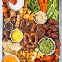 an assortment of snack foods on a tray like pretzels, chicken wings, cheese, veggie sticks, and tortilla chips scattered around dips like guacamole, mustard, and ranch