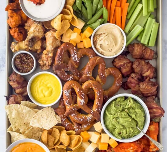 an assortment of snack foods on a tray like pretzels, chicken wings, cheese, veggie sticks, and tortilla chips scattered around dips like guacamole, mustard, and ranch