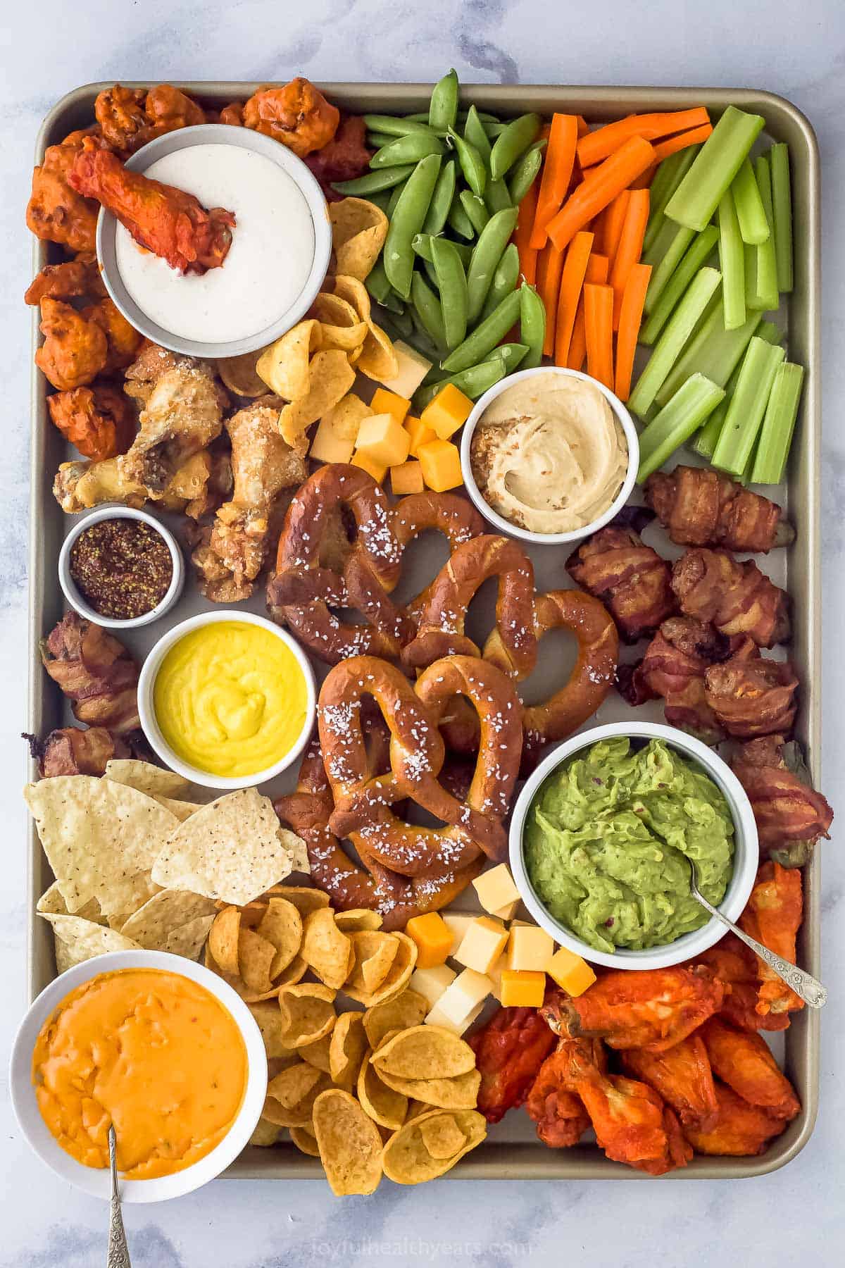 an assortment of snack foods on a tray like pretzels, chicken wings, cheese, veggie sticks, and tortilla chips scattered around dips like guacamole, mustard, and ranch