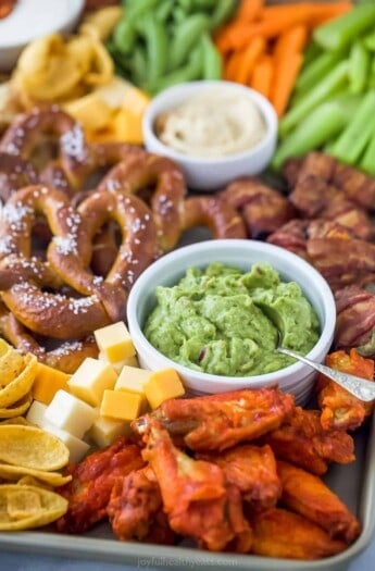 an assortment of snacks including a small bowl of guacamole, pretzels, cheese cubes, and chicken wings