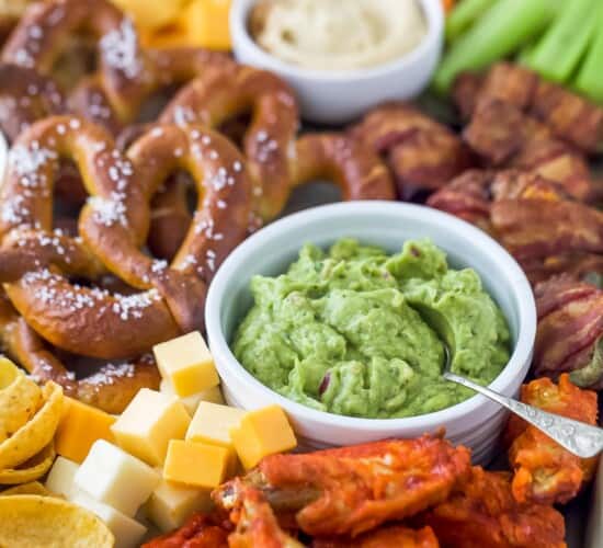 an assortment of snacks including a small bowl of guacamole, pretzels, cheese cubes, and chicken wings