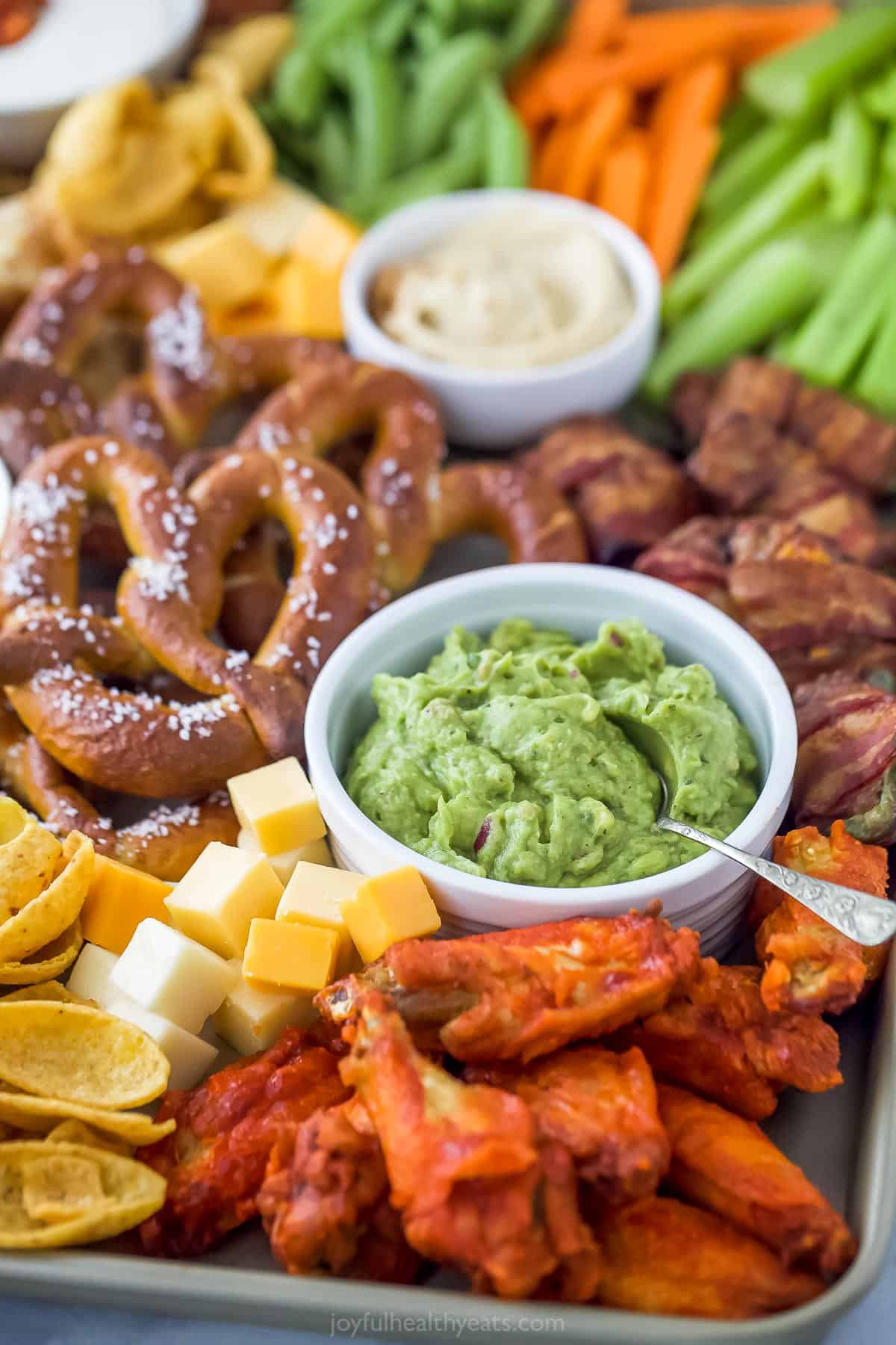 an assortment of snacks including a small bowl of guacamole, pretzels, cheese cubes, and chicken wings