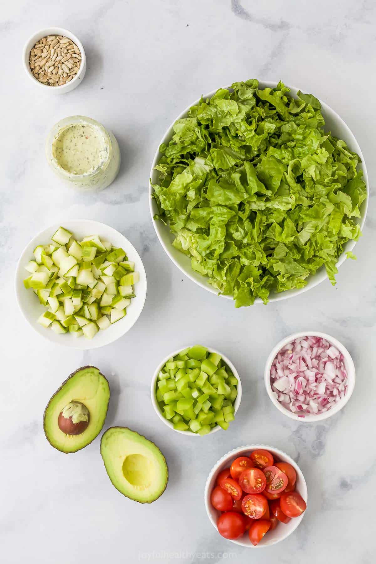 An avocado, a jar of dressing, a dish full of sunflower seeds and the rest of the ingredients in bowls on a kitchen countertop